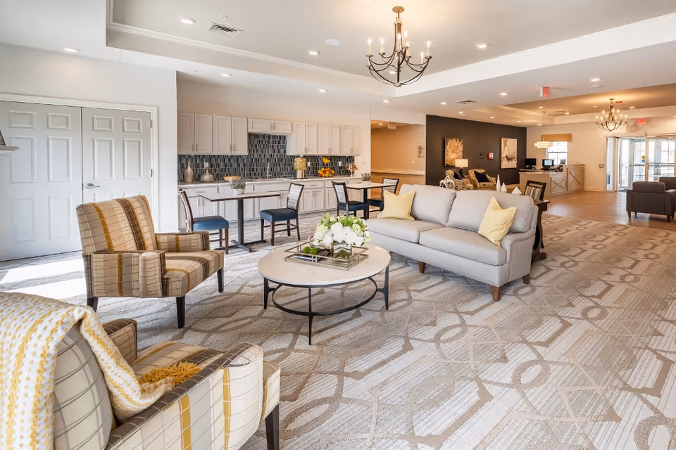 A spacious and well-lit common area in a senior living facility featuring a light gray sofa with yellow pillows, two plaid armchairs, a round coffee table with a floral centerpiece, and a kitchenette with white cabinets and a tiled backsplash. The room has patterned carpet flooring, chandeliers, and multiple seating areas with tables and chairs.