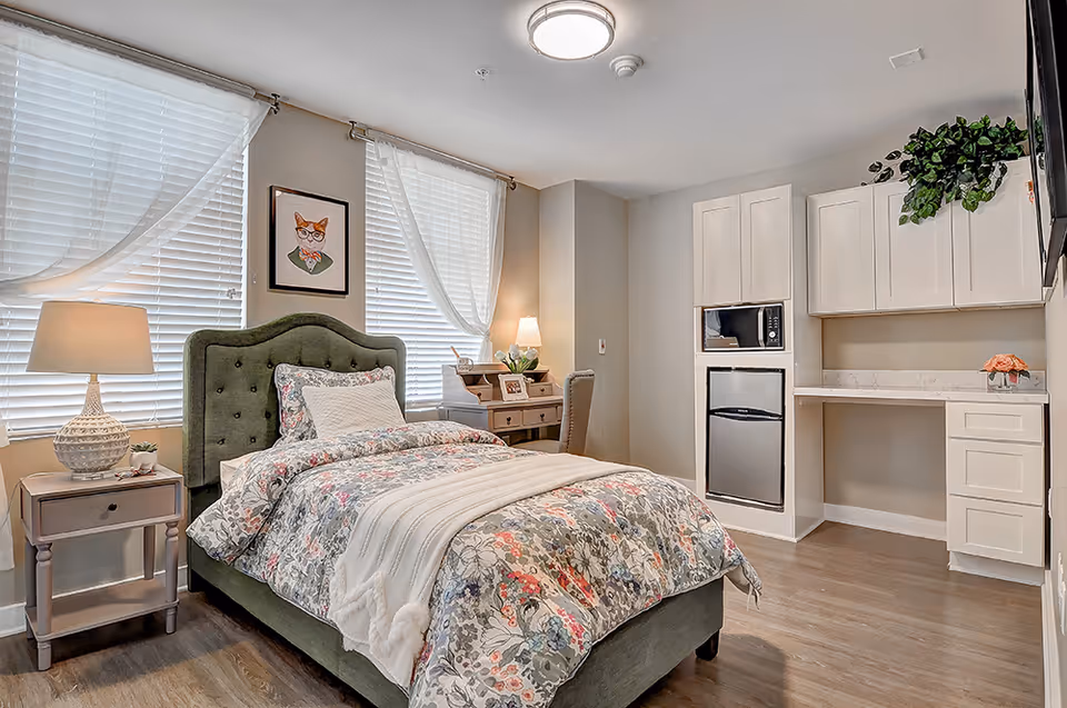 A cozy bedroom with a green upholstered bed featuring floral bedding and a white throw blanket. There are two windows with white blinds and sheer curtains, a nightstand with a lamp and small plant, a desk with a chair and lamp, and a kitchenette area with white cabinets, a microwave, and a mini fridge. A framed picture of a cat wearing glasses and a suit hangs on the wall between the windows.