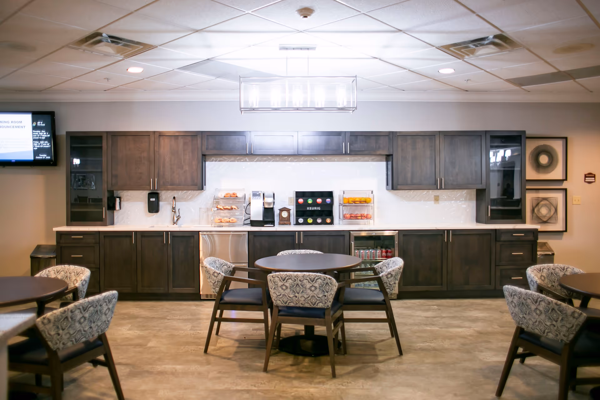 A modern dining area in a senior living facility with round tables and cushioned chairs. The back wall features dark wood cabinets, a coffee machine, a Keurig beverage station, and display cases with pastries and fruit. A flat-screen TV is mounted on the left wall, and framed artwork is on the right wall. The floor is a light-colored tile, and a rectangular light fixture hangs from the ceiling.