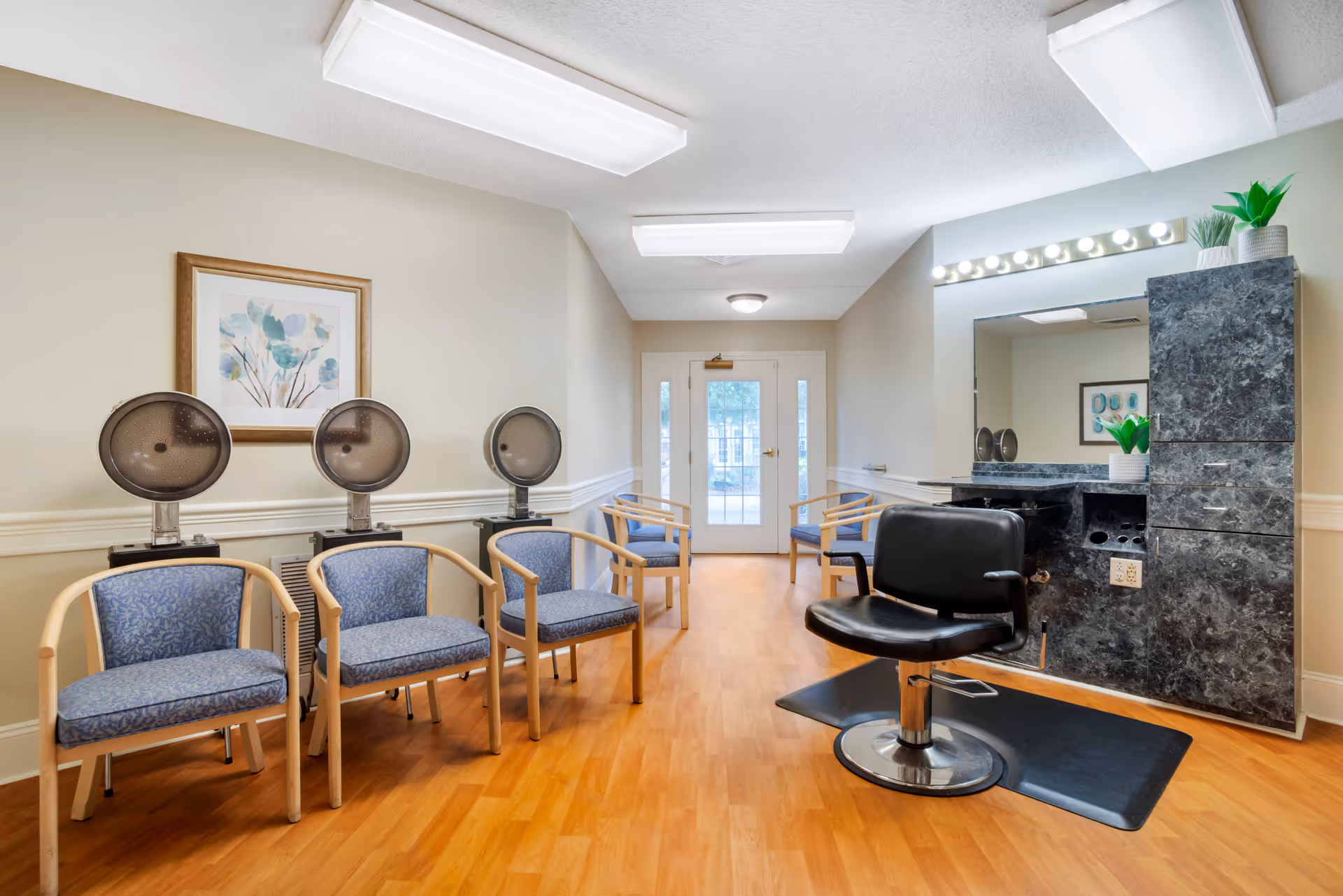 Interior view of a hair salon area in a senior living facility with three blue cushioned chairs under hair dryers along the left wall, a black salon chair in front of a large mirror with a black marble cabinet, and a door with glass panels at the end of the room.