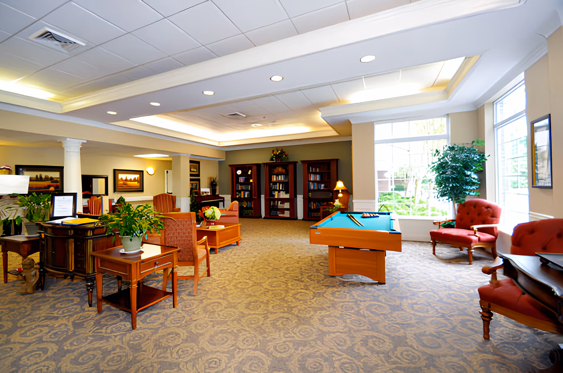 Spacious senior living common room with seating, a pool table, bookshelves, and large windows.