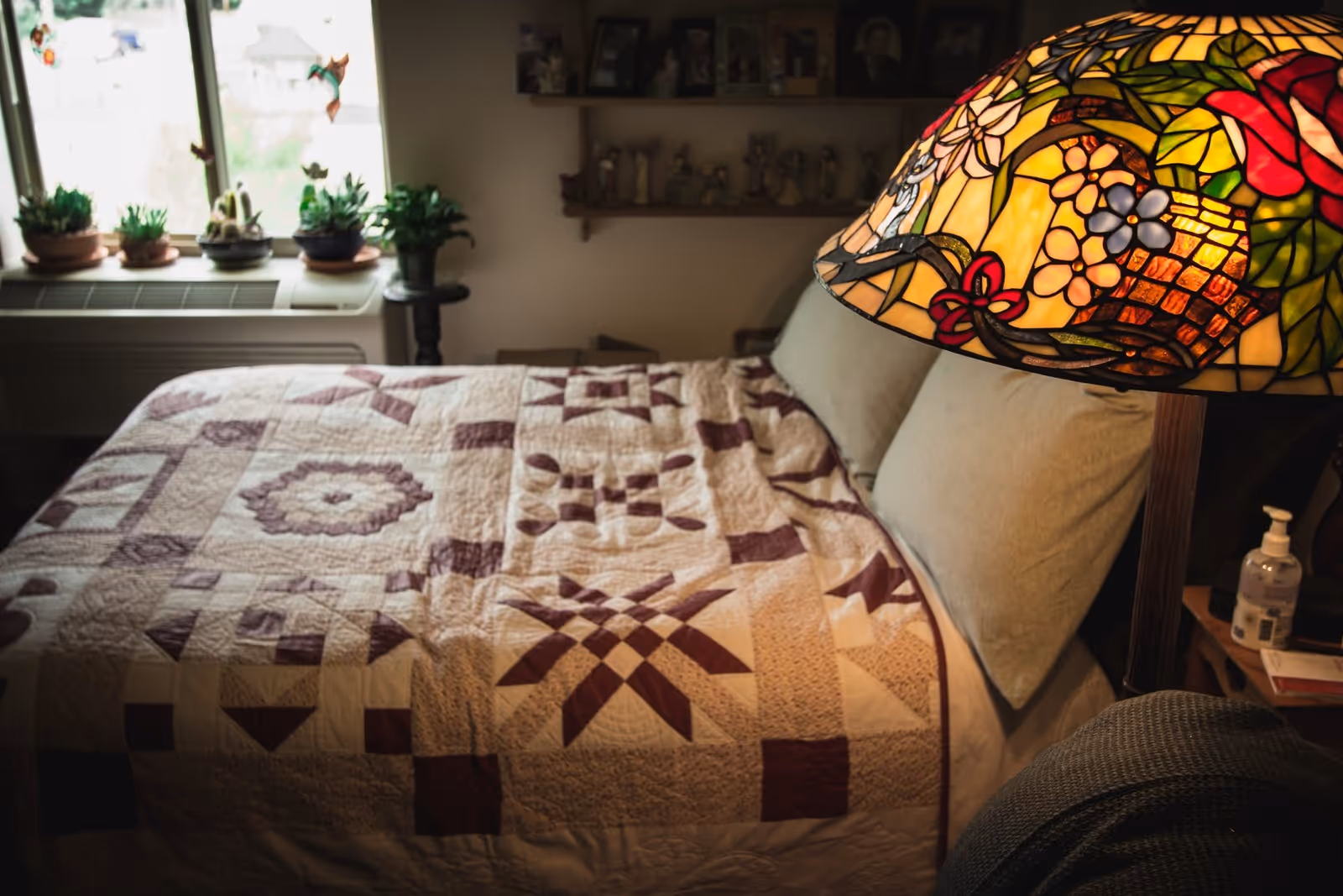 A cozy bedroom with a bed covered in a quilt featuring various geometric patterns in shades of beige and brown. Two light green pillows rest against the headboard. A colorful stained glass lamp with floral designs is on a bedside table, which also holds a bottle of hand sanitizer and some books. In the background, there is a window with several potted plants on the windowsill and a shelf with decorative figurines on the wall.