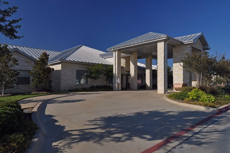 Exterior view of a single-story building with light-colored stone walls and a metal roof under a clear blue sky. The entrance features a covered drop-off area supported by stone pillars, surrounded by landscaped greenery and trees.