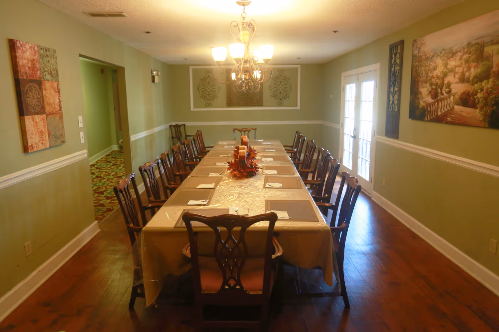 Long dining room with a table set for many guests, chairs along both sides, a chandelier, and artwork on green walls.