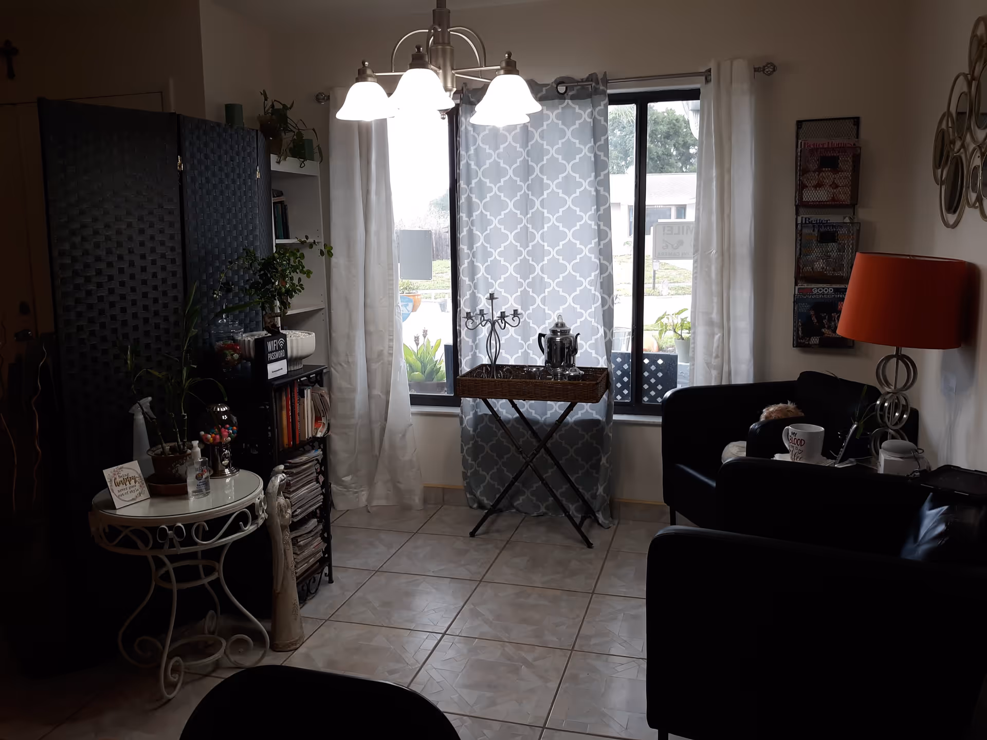 A cozy living room area with two black armchairs, a small round white table with plants and decorative items, a black folding screen, a bookshelf filled with books and magazines, a window with patterned and white curtains, a small table with a silver coffee pot and candle holder, and a red lamp on a side table. The floor is tiled and the room is softly lit by a ceiling light fixture.