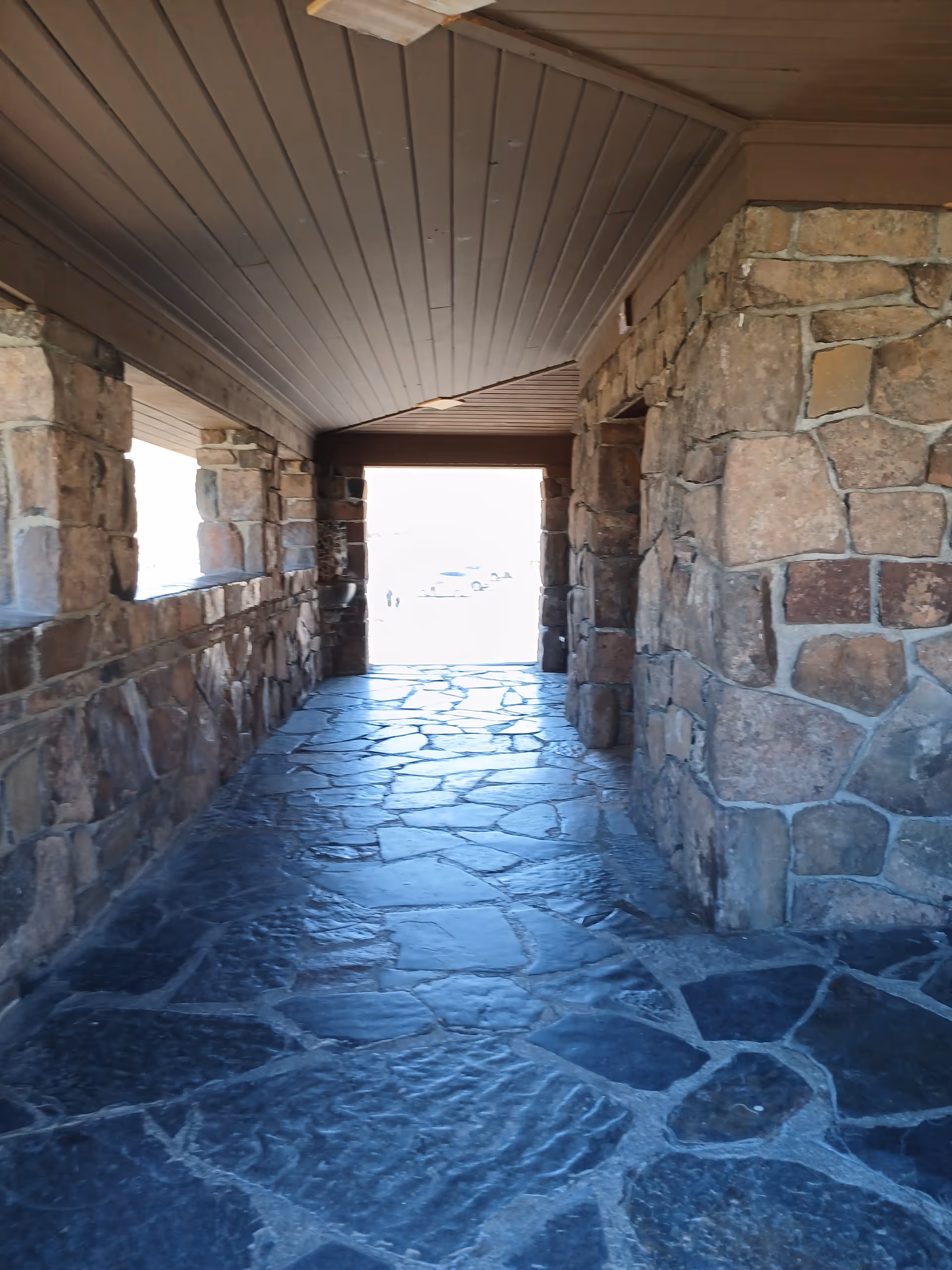 Covered stone passageway with a flagstone floor and stone walls opening to bright daylight.