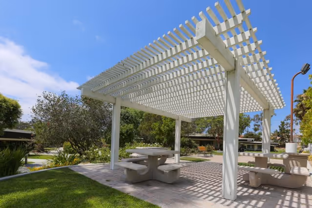 Outdoor seating area with white pergola providing shade over concrete picnic tables and benches, surrounded by green grass, trees, and shrubs under a clear blue sky.