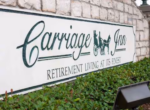 Exterior sign for Carriage Inn Katy retirement living beside a stone wall and greenery.
