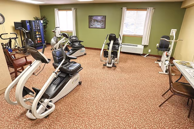 A fitness room with exercise equipment including stationary bikes and other workout machines. The room has green walls, two windows with white curtains, a vending machine, a chair, and a table with a chair. The floor is covered with a patterned carpet.