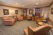A cozy sitting area in a senior living facility with several upholstered armchairs arranged around small wooden tables. The room features carpeted flooring, framed artwork on the walls, built-in bookshelves filled with books, and warm lighting from table lamps.