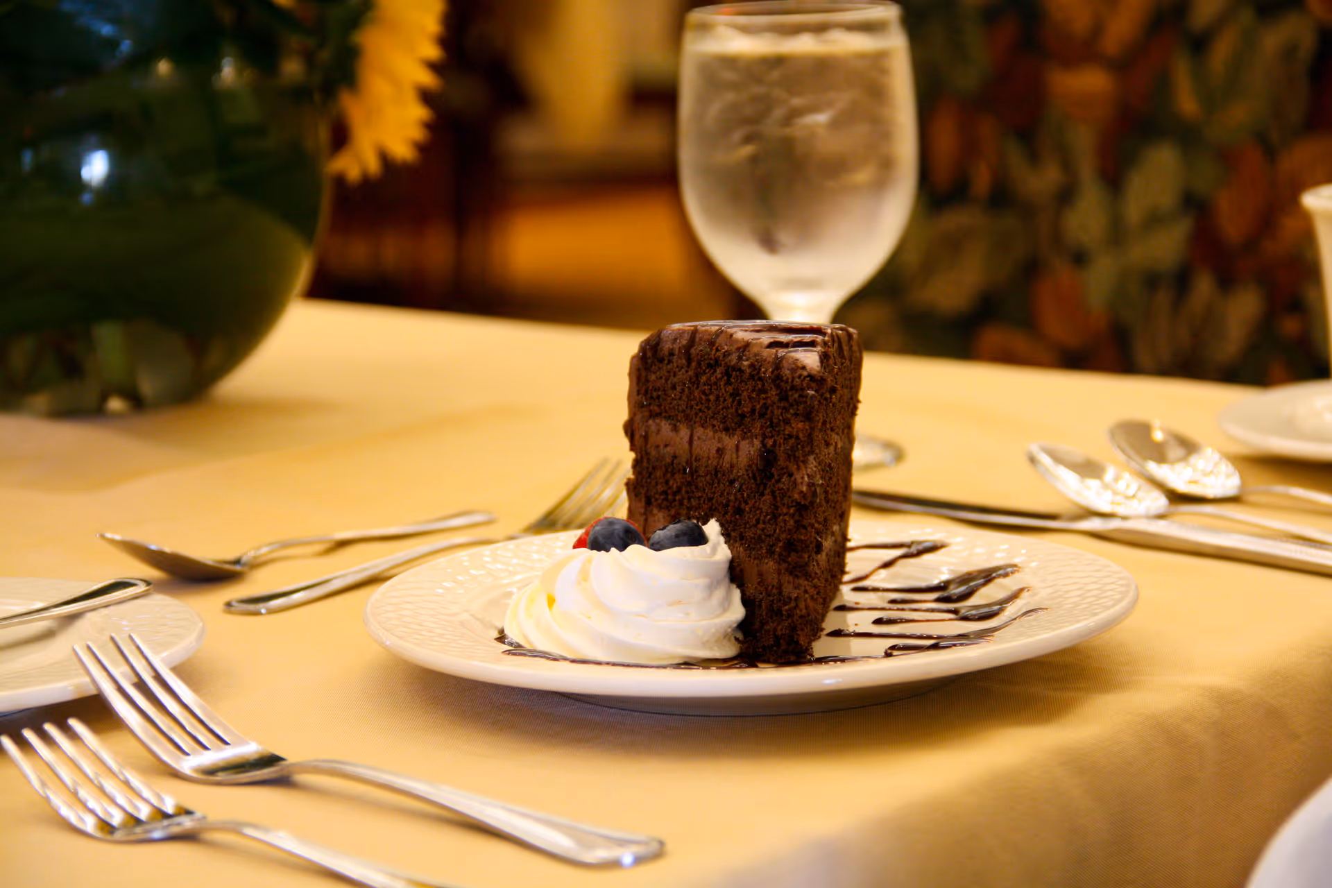 A slice of chocolate cake with whipped cream and blueberries on a white plate drizzled with chocolate sauce, placed on a table set with silverware and a glass of water in the background.