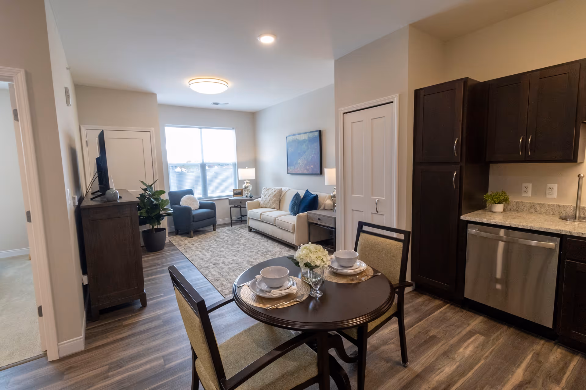 A bright and modern living space featuring a small round dining table set for two with white bowls, plates, and glasses. The room includes a beige sofa with blue and beige pillows, a blue armchair, a TV on a wooden stand, and dark wood kitchen cabinets with a granite countertop and stainless steel dishwasher. A large window lets in natural light, and the floor is covered with wood-style flooring and a patterned area rug.
