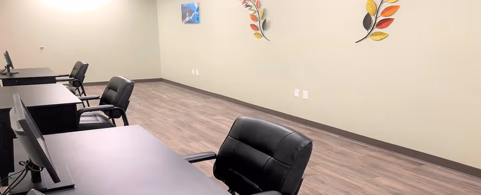 A room with several black office chairs and desks arranged in a row along a wall. The floor has wood-like vinyl flooring, and the walls are painted light green. Two decorative metal leaf wall hangings and a small framed picture are visible on the wall.