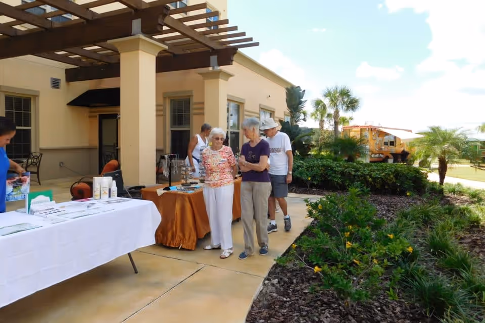 Several elderly people standing and walking near tables with informational materials set up outside a beige building with a pergola. There are plants and palm trees along the walkway, and a food truck is visible in the background under a partly cloudy sky.