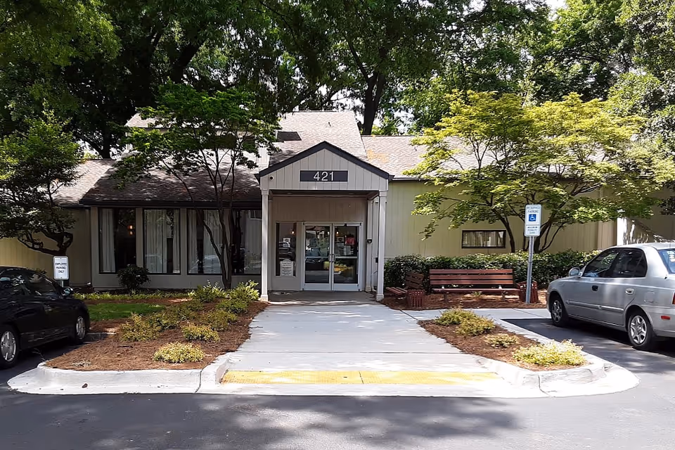 Front entrance of a single-story building with the number 421 above the door. The building is surrounded by trees and shrubs, with a sidewalk leading to the entrance. There are two parked cars on either side of the entrance and a bench to the right near a handicap parking sign.
