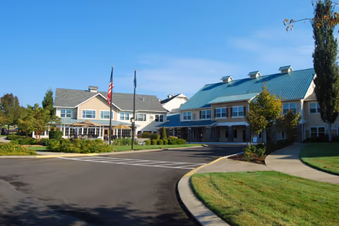 Exterior view of a senior living facility with two main buildings, one with a blue roof and the other with a gray roof. There are two flagpoles with the American flag and another flag in front of the buildings. The area is landscaped with green grass, trees, and shrubs under a clear blue sky.