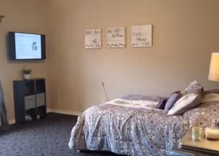 A bedroom in a senior living community featuring a bed with patterned bedding and multiple pillows. On the wall above the bed are three decorative framed quotes. To the left, there is a small shelving unit with a plant on top and a wall-mounted TV above it. A lamp is visible on a bedside table to the right.