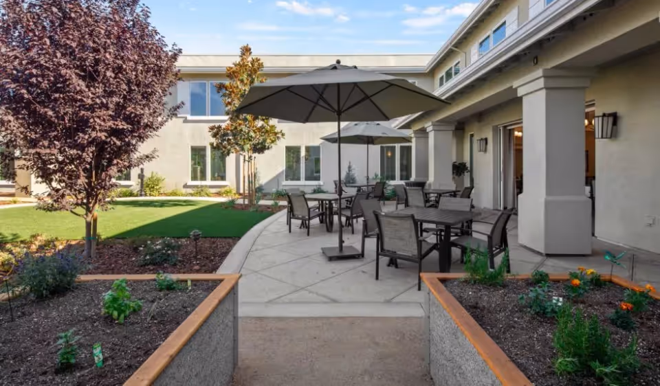 Outdoor patio area at Oakmont of Lodi with multiple tables and chairs under large umbrellas, surrounded by garden beds with plants and trees, adjacent to a building with large windows and columns.