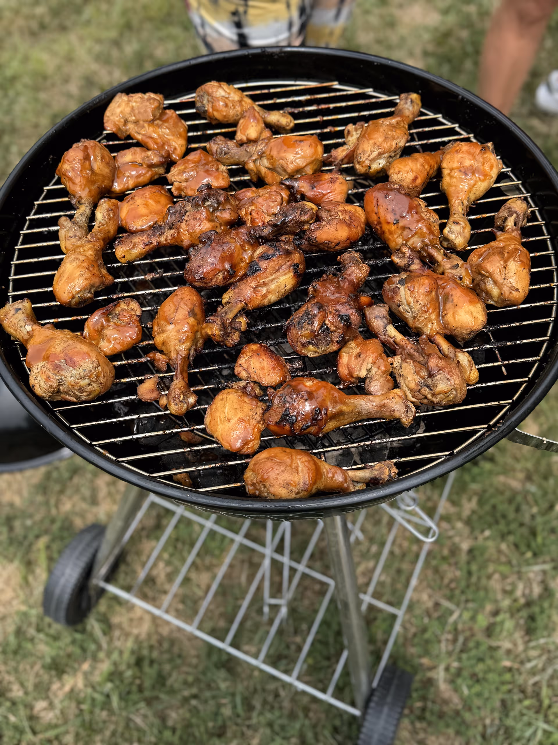 Charcoal grill filled with many grilled chicken drumsticks outdoors on grass.
