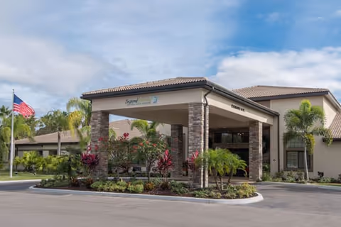 Entrance porte-cochere of a single-story senior living building with stone columns, landscaped beds, palm trees and an American flag.
