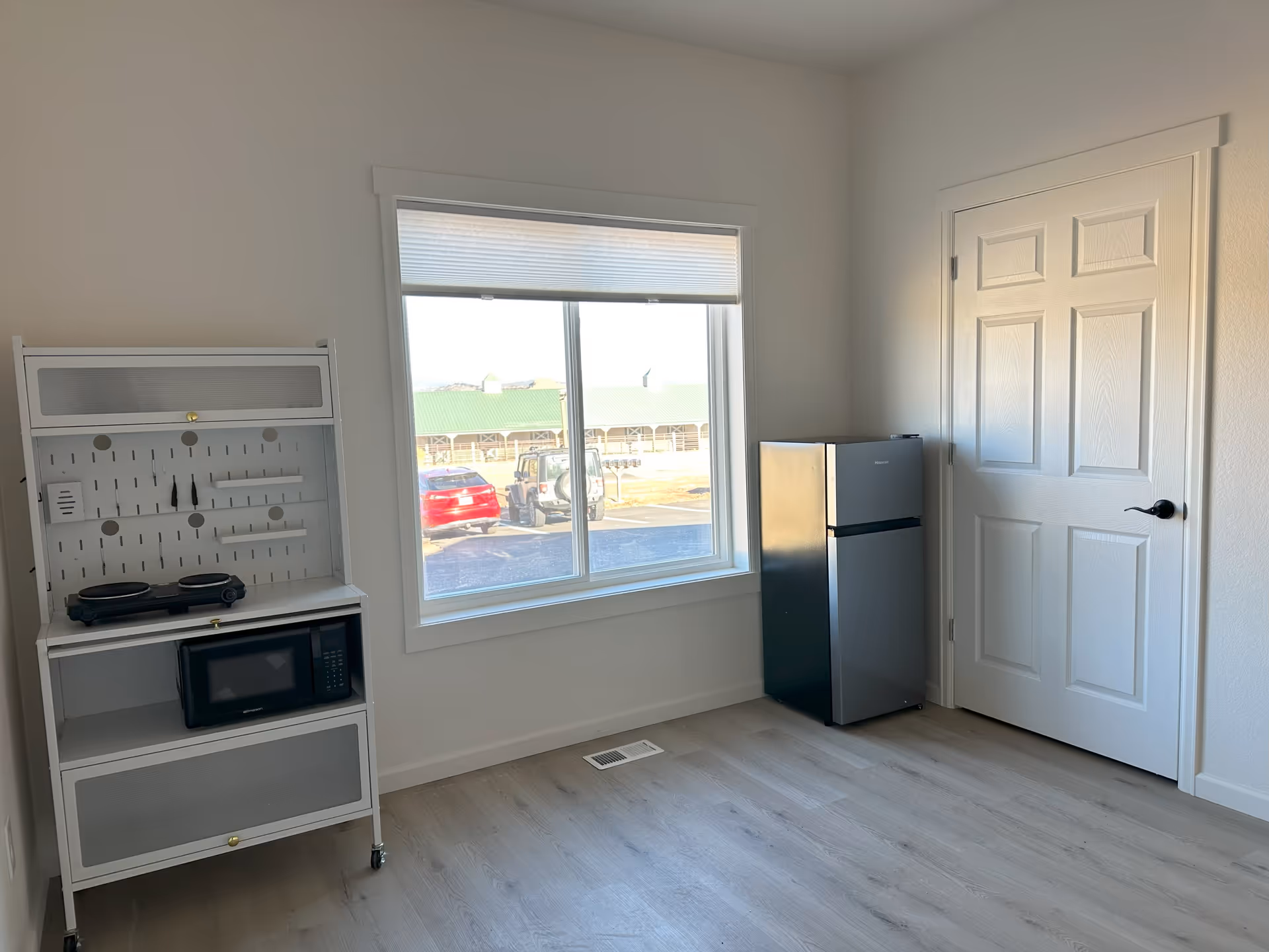 Small bright room with a window, a mini fridge, a microwave cart and a closed white door.