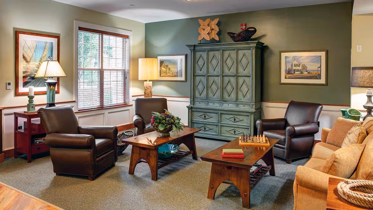 A cozy living room with two dark brown leather armchairs, a beige sofa, two wooden coffee tables with books and a chessboard, a green cabinet with decorative items on top, framed artwork on the walls, and two table lamps providing warm lighting.