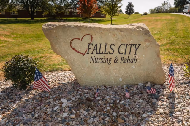 A large stone with the inscription 'Falls City Nursing & Rehab' and a red heart symbol, placed on a bed of small rocks with several small American flags around it. In the background, there is a grassy area with trees and a building partially visible.