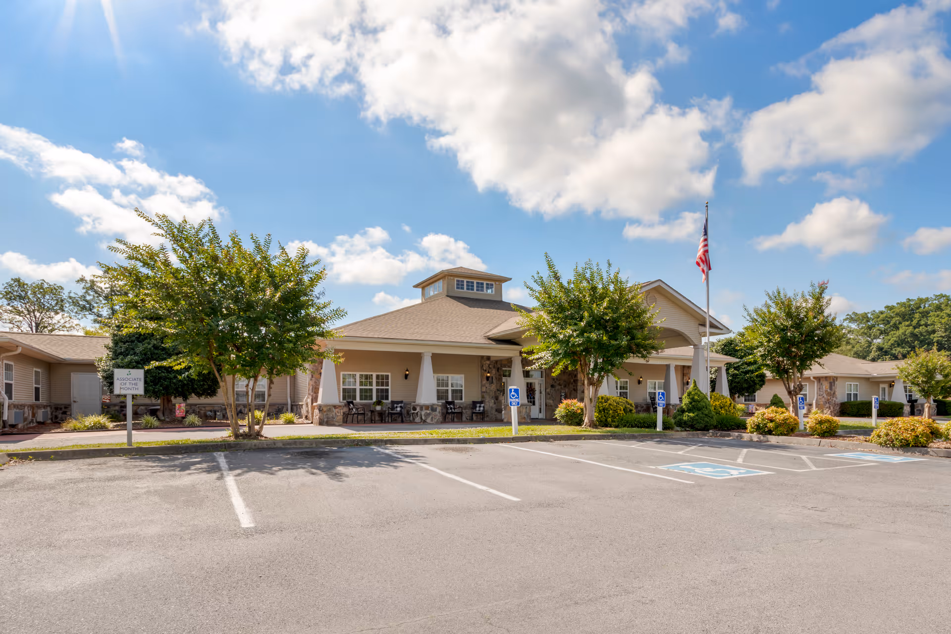 Exterior view of Brookdale Sevierville senior living facility on a sunny day with a clear blue sky and scattered clouds. The building features a covered entrance with stone pillars, several windows, and a flagpole with an American flag. There are trees and shrubs around the building and a parking lot with marked handicap spaces in the foreground.