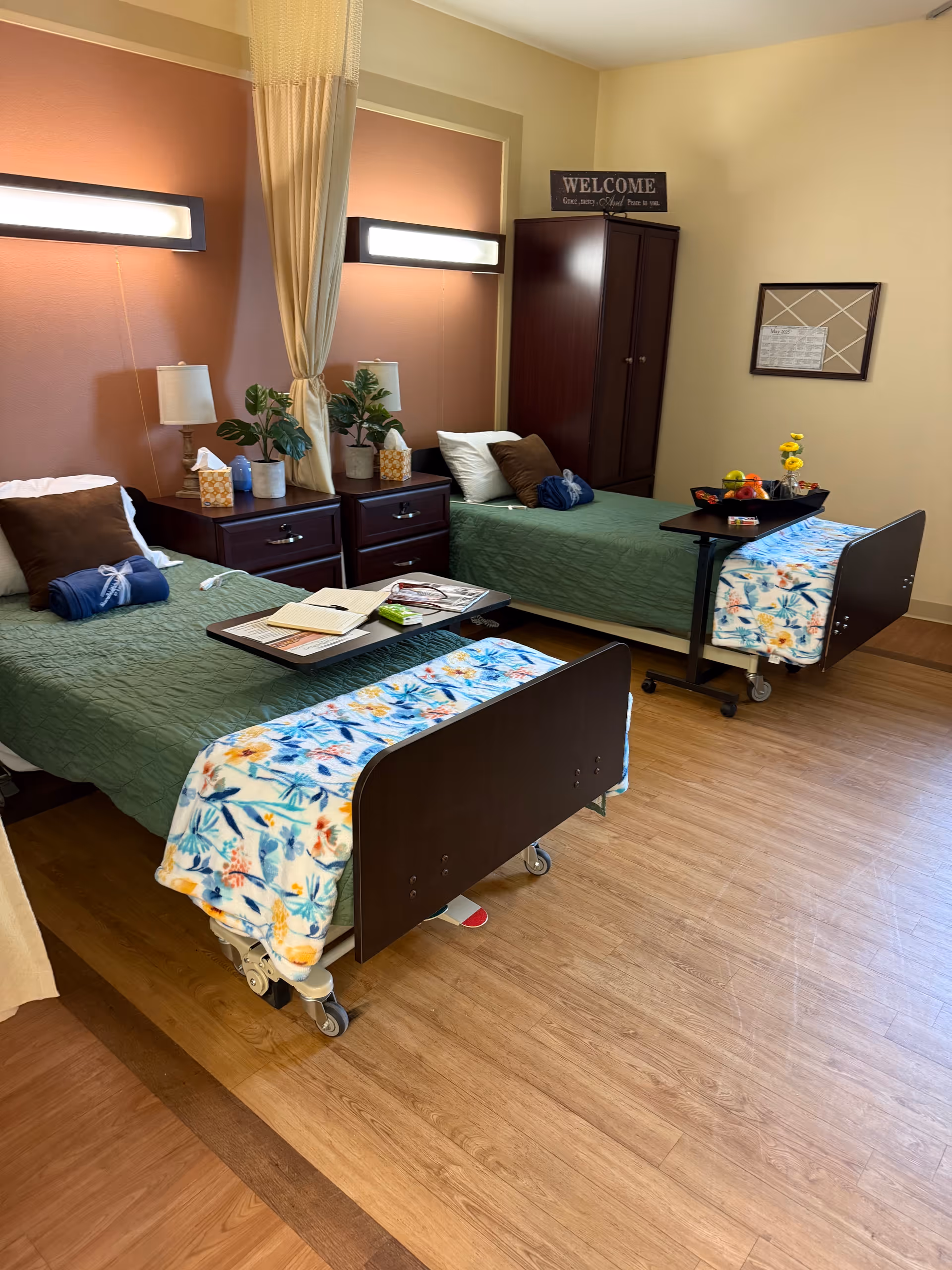 A shared bedroom in a senior living facility with two hospital-style beds, each with green bedspreads and floral blankets at the foot. Each bed has pillows and a rolled towel. Between the beds are two dark wooden nightstands with lamps, plants, and tissue boxes. A curtain hangs between the beds for privacy. A dark wooden wardrobe stands against the wall with a 'WELCOME' sign above it. One bed has a tray table with an open book and reading glasses, while the other has a tray with fruit and a small vase with yellow flowers. The room has wood flooring and warm beige walls.