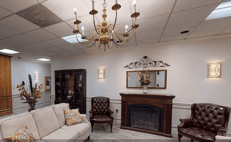 A cozy living room area with a beige sofa adorned with patterned pillows, two dark brown leather armchairs, a wooden fireplace with a decorative mirror and floral arrangement above it, a wooden cabinet with glass doors, and a gold chandelier hanging from the ceiling.