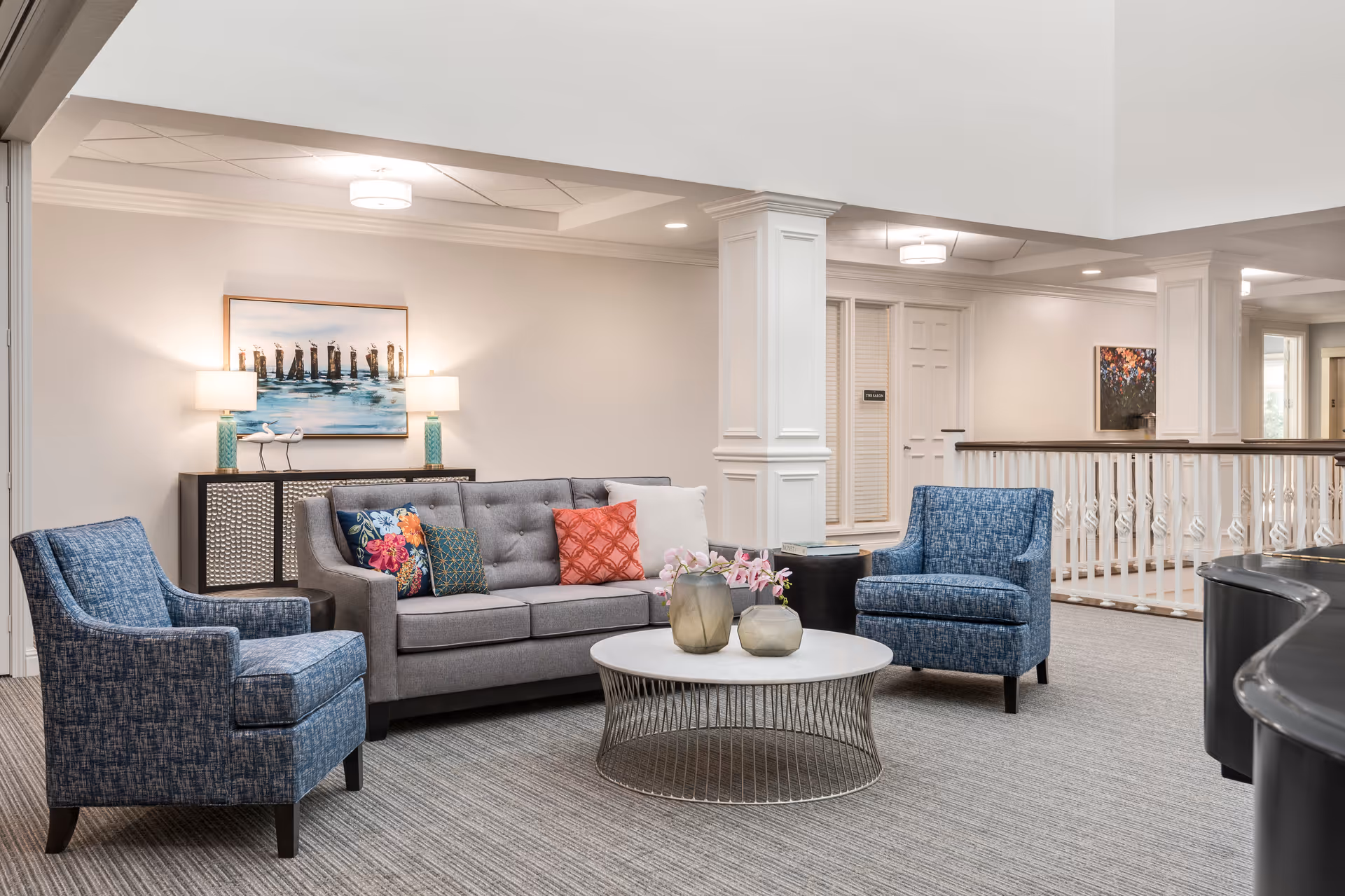 A bright and spacious living room area with a gray sofa adorned with colorful pillows, two blue upholstered armchairs, and a round coffee table with decorative vases. The room features white walls, a carpeted floor, a sideboard with two lamps and a painting above it, and white columns with a railing in the background.