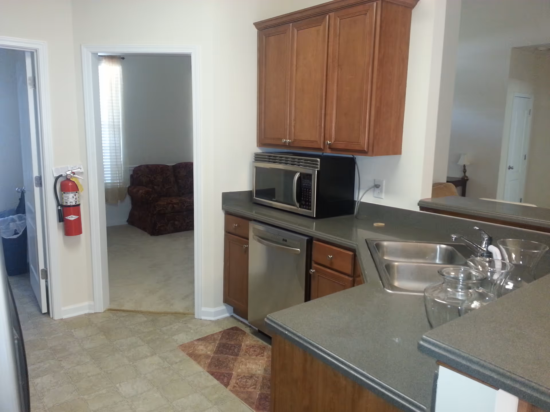 Interior view of a kitchen area with wooden cabinets, a microwave, a dishwasher, a double sink, and glass containers on the counter. There is a doorway leading to a room with a patterned sofa and a window with blinds. A fire extinguisher is mounted on the wall near the doorway.