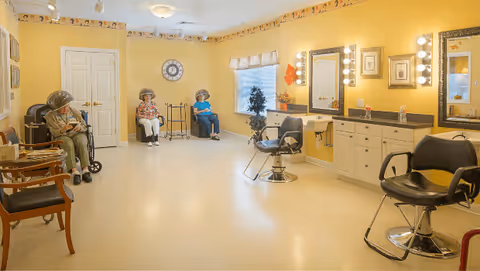 A bright yellow room in a senior living facility with three elderly women sitting under hair dryers along the left wall. The room has salon chairs, mirrors with lights, cabinets, and a window with a valance. The walls are decorated with a border featuring small images near the ceiling.
