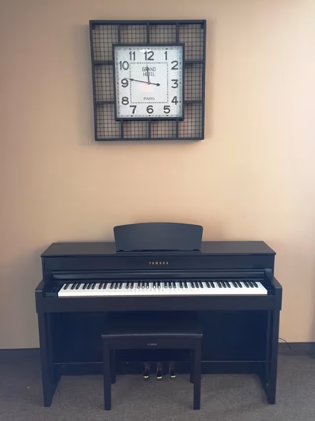 A black Yamaha digital piano with a bench sits beneath a square wall clock on a beige wall.