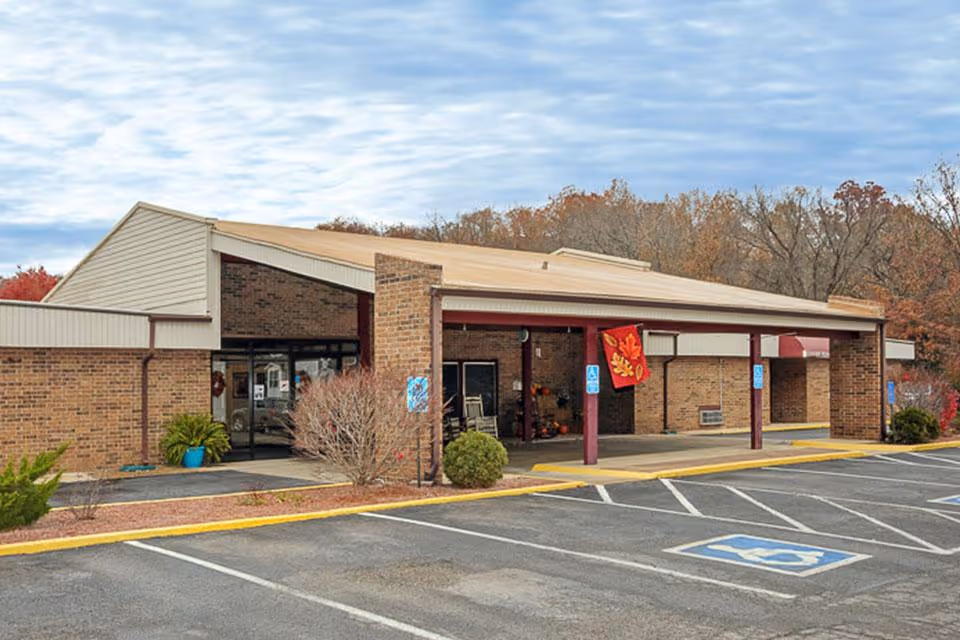 Exterior view of a single-story brick building with a covered entrance and several handicap parking spaces in front. The building has a beige roof and maroon support beams. There are some bushes and small trees around the entrance, and a red flag with autumn leaves is hanging near the doorway. The sky is partly cloudy.