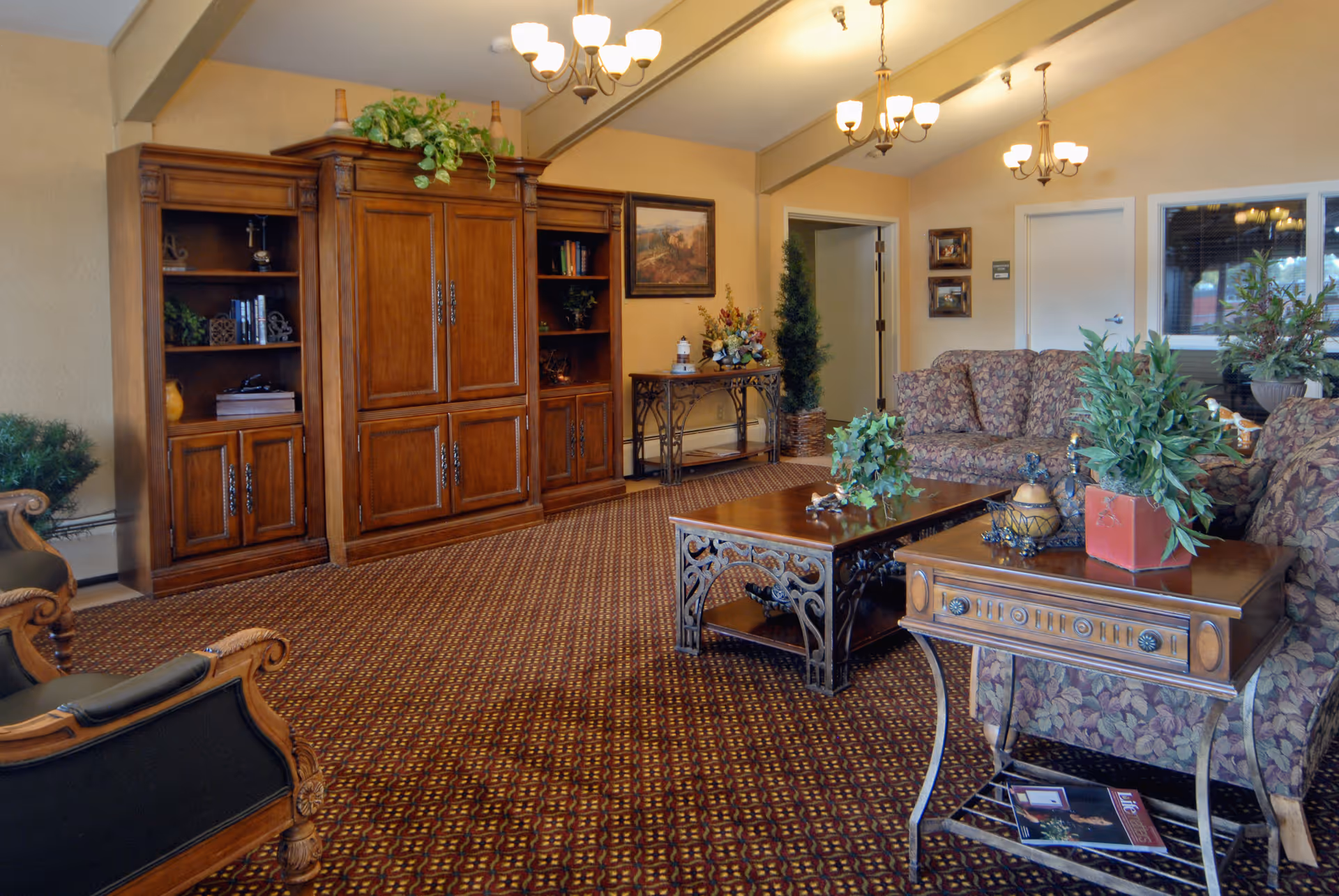 A cozy living room area in a senior living facility with patterned carpet, floral upholstered sofas, wooden coffee and side tables with decorative plants, a large wooden entertainment center, and warm lighting from chandeliers.