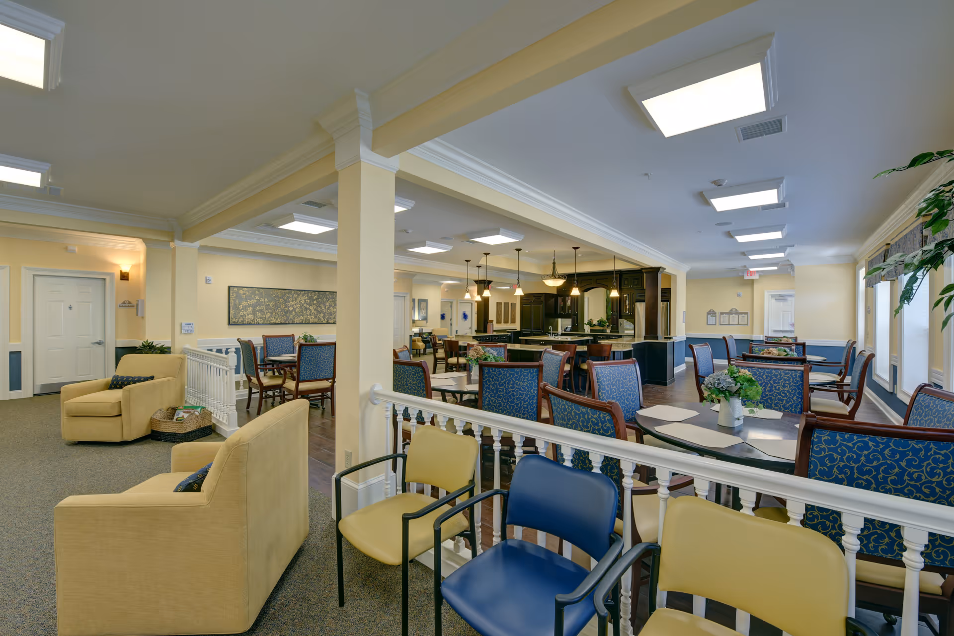 Interior view of a senior living facility common area with multiple seating arrangements including yellow armchairs, blue and yellow chairs, and dining tables with blue upholstered chairs. The room has soft yellow walls, white trim, and ceiling lights. In the background, there is a kitchen area with dark cabinetry and pendant lighting.
