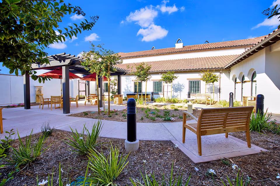 Outdoor courtyard area at Ridgeview Health Center featuring wooden benches, small trees, plants, a pergola with red umbrellas, and a clear blue sky with a few clouds.