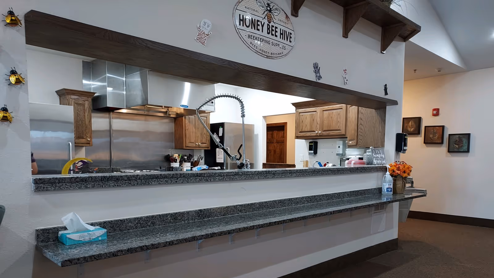 Interior view of a kitchen area with a long granite countertop and a large pass-through window. The kitchen has wooden cabinets, a stainless steel refrigerator, and a commercial-style faucet. Above the pass-through window is a decorative sign that reads 'Honey Bee Hive Beekeeping Supplies' with small bee decorations on the wall. On the countertop, there is a box of tissues, a bottle of hand sanitizer, a vase with orange flowers, and a small sign that says 'Live Simply Grateful'.