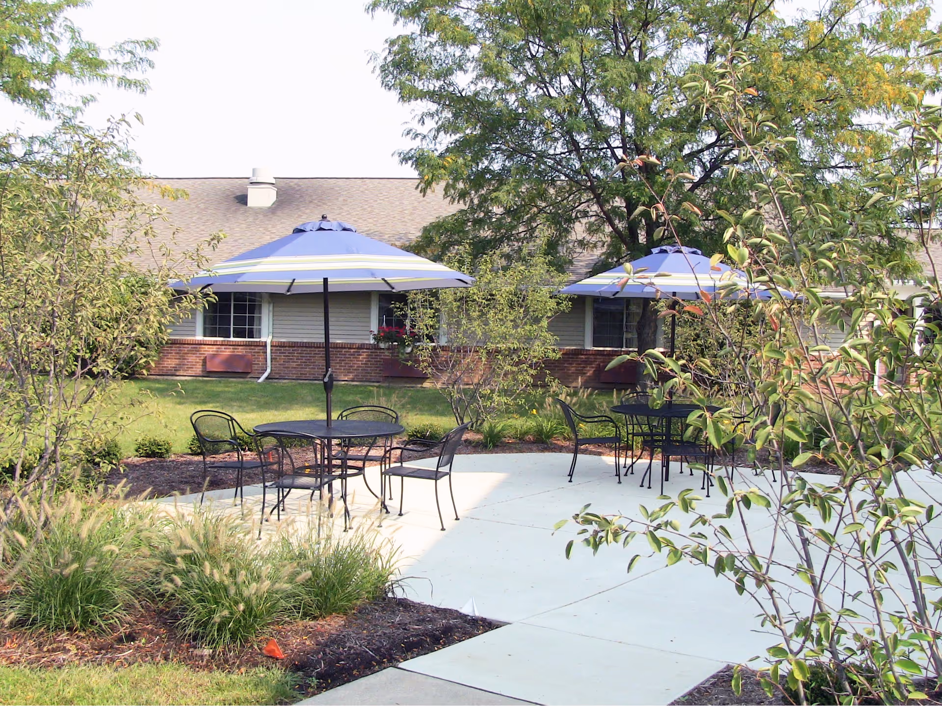Outdoor patio area with two black metal tables and chairs, each table shaded by a blue and white striped umbrella. The patio is surrounded by green grass, bushes, and trees, with a single-story building with brick and siding in the background.