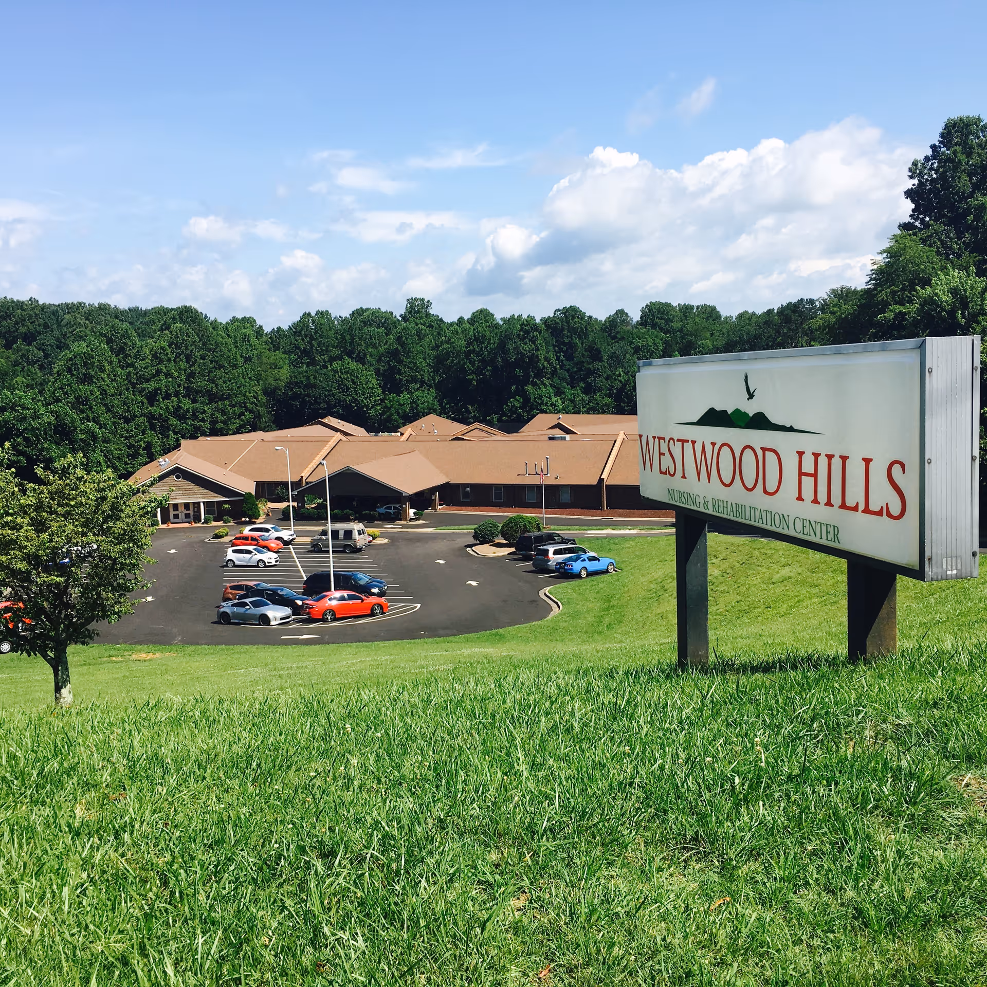 View of Westwood Hills Nursing and Rehabilitation Center building with a parking lot in front, surrounded by green grass and trees under a partly cloudy blue sky. A large sign with the facility's name is prominently displayed on the right side of the image.