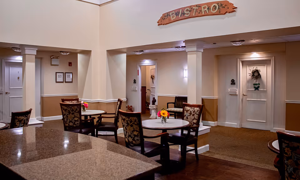 Interior view of a senior living facility dining area with round tables and floral upholstered chairs. Each table has a small vase with colorful flowers. The walls are painted beige and white, and there is a sign above that reads 'BISTRO'. Several doors and a bench are visible in the background.