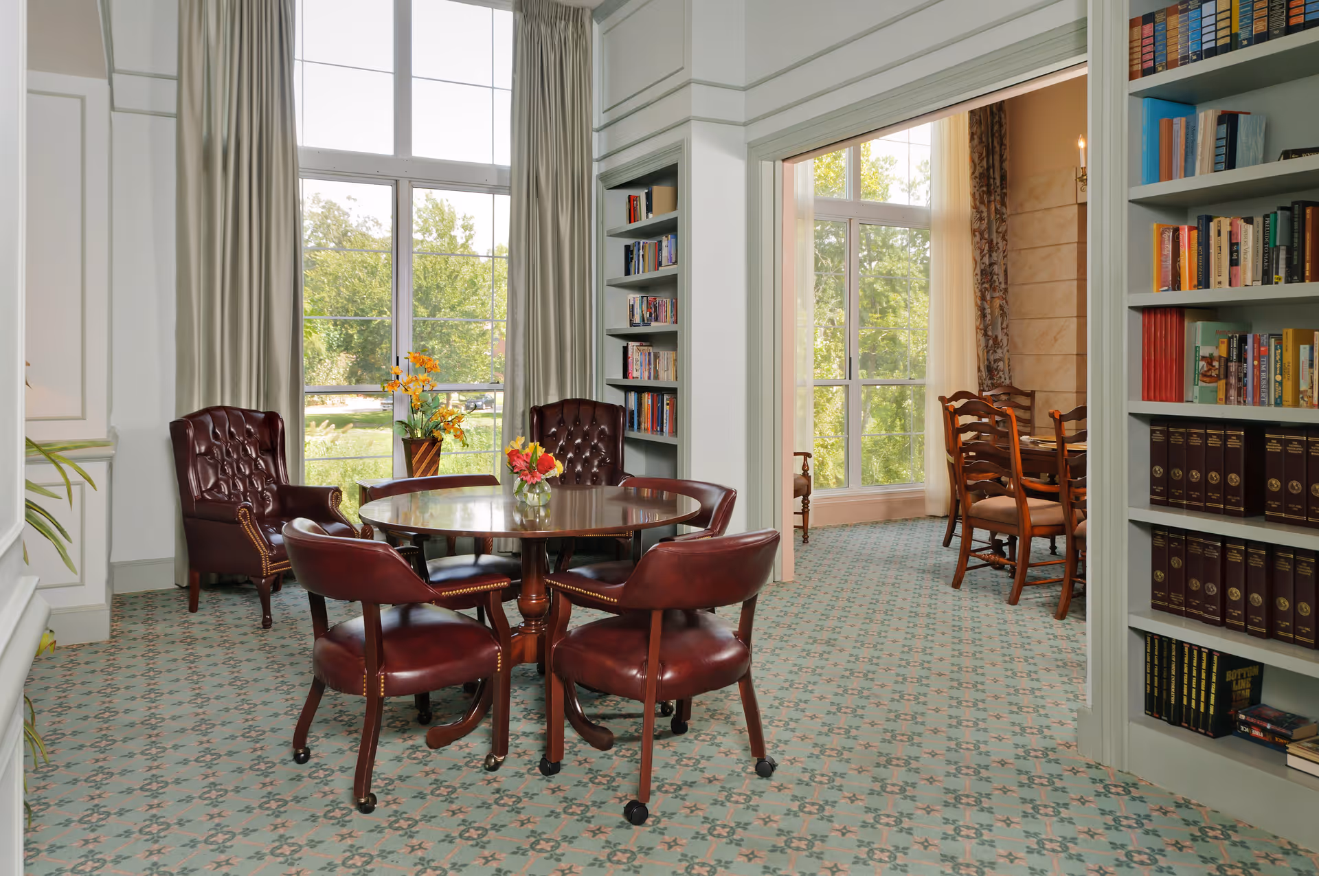 A cozy interior room with a round wooden table surrounded by four leather chairs on casters. The room has large windows with beige curtains letting in natural light and a view of greenery outside. Built-in bookshelves filled with books flank the doorway leading to another room with a wooden dining table and chairs. The carpet has a patterned design in soft colors.