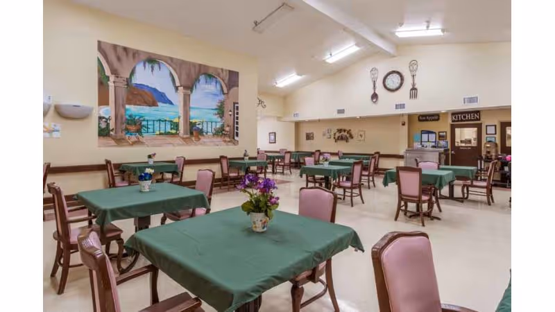 A spacious dining room with multiple tables covered in green tablecloths and surrounded by wooden chairs with pink cushions. The walls are decorated with a large mural depicting an ocean view through arches, a clock, and oversized decorative utensils. There is a door labeled 'KITCHEN' in the background.