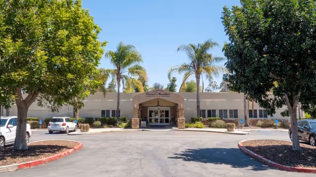 Front exterior view of Carmel Mountain Rehabilitation And Healthcare Center with a driveway, parking spaces, palm trees, and other greenery under a clear blue sky.