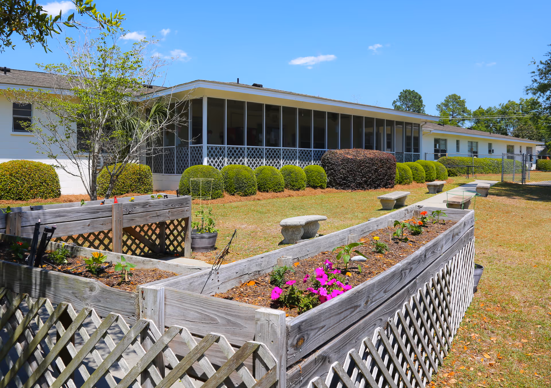 Raised wooden planter boxes with flowers and stone benches on a lawn in front of a single-story building with a screened porch.