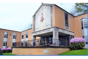 Exterior view of Vincentian De Marillac facility showing a brick building with large windows and an entrance area with a covered porch. There is a religious emblem above the entrance and well-maintained landscaping with green grass and purple flowering bushes.