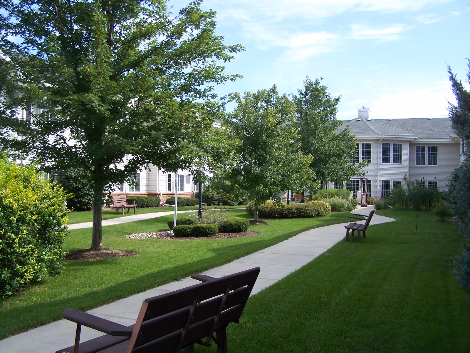 A landscaped outdoor courtyard area with a paved walking path, green grass, several trees, bushes, and multiple wooden benches. In the background, there is a two-story building with many windows under a partly cloudy sky.