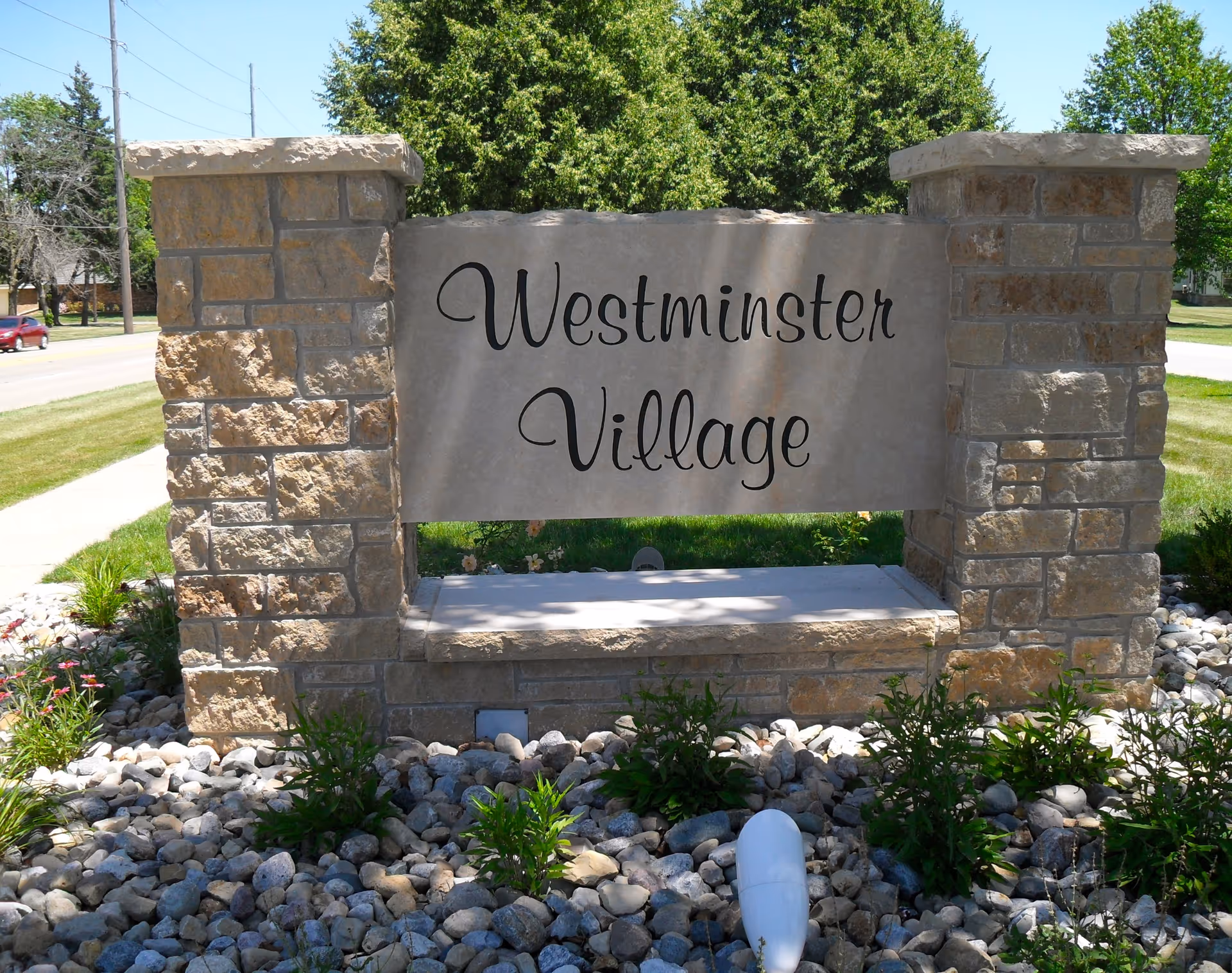 Stone sign with the text 'Westminster Village' surrounded by landscaping with rocks and plants, located near a sidewalk and street with trees in the background.