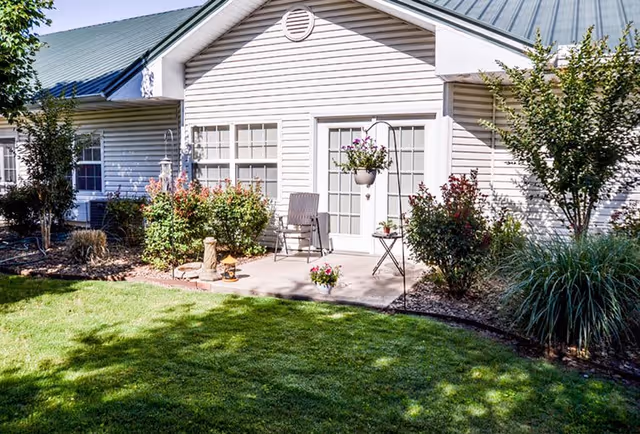 A sunny outdoor patio area of a residential building with white siding and a metal roof. The patio has a small table, two chairs, potted plants, and hanging flower baskets. Surrounding the patio are neatly trimmed bushes, small trees, and a well-maintained green lawn.